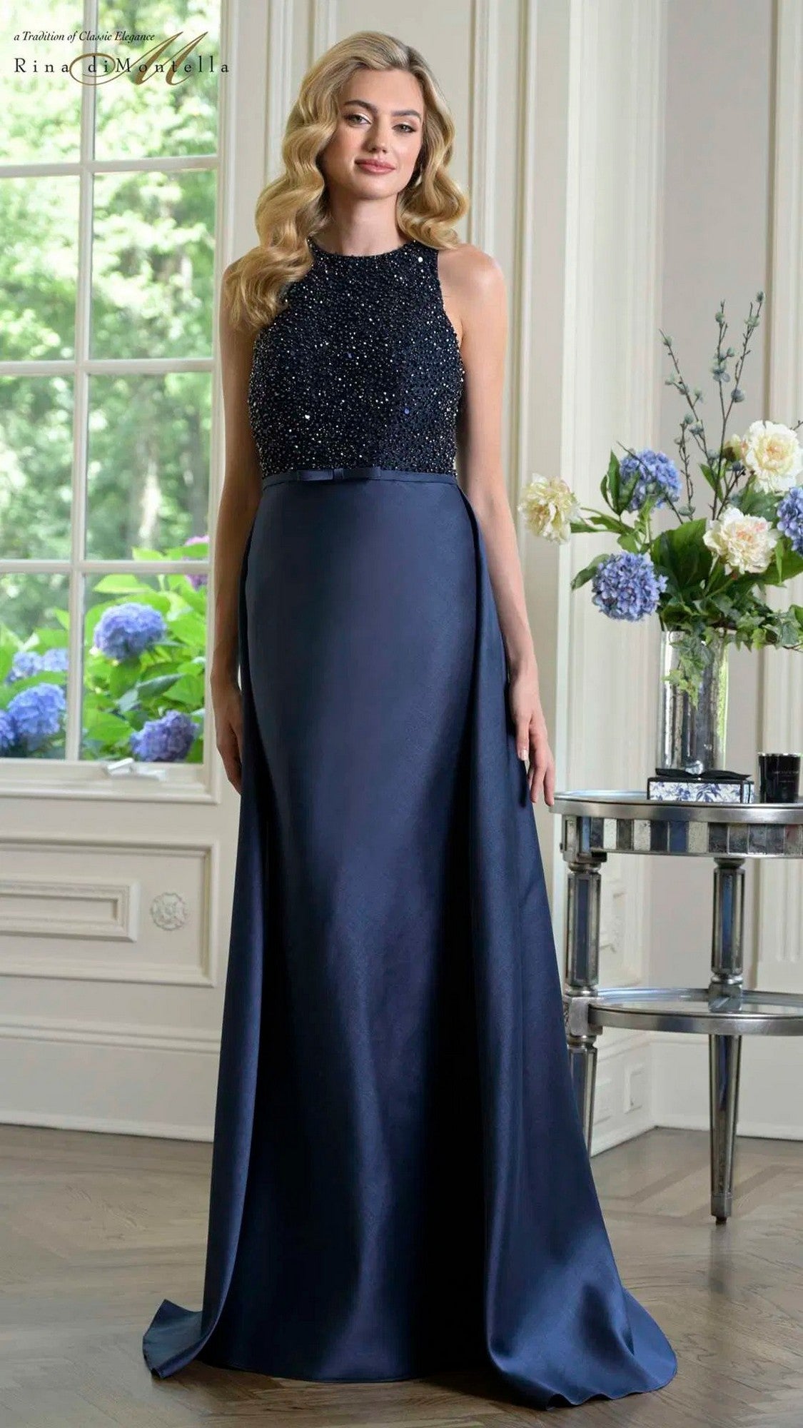 A woman wearing the Rina Di Montella RD3404 long formal navy dress with a beaded bodice and floor-length skirt stands indoors near a table adorned with flower arrangements.
